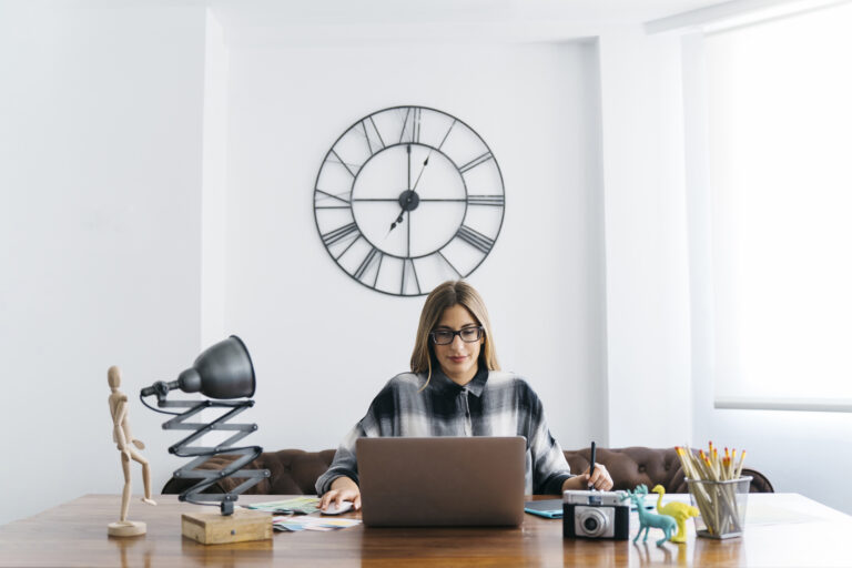 Mujer trabajando en su escritorio, con un gran reloj a sus espaldas en la pared, haciendo alusión de la importancia de la flexibilidad horaria en el trabajo en la actualidad.